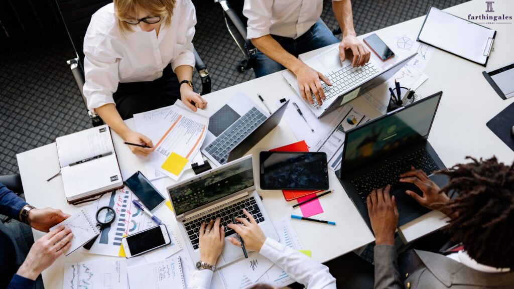 Overhead view of a diverse professional team sitting around a white conference table collaborating on laptops and reviewing paper documents.