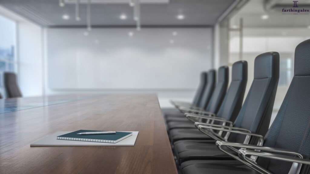 Low-angle shot of a bright, modern boardroom featuring a row of sleek black leather chairs lined up alongside a polished wooden conference table, with a notebook and pen sitting in the foreground.